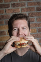 White middle aged man bites into hamburger with ketchup on a sesame bun looking to camera
