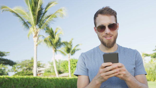Smartphone man sms texting using app on smart phone outdoors in summer. Handsome young casual man using mobile cell phone smiling happy wearing sunglasses. Urban male hipster. RED EPIC 90 FPS.