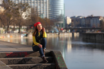 Beautiful Asian woman in yellow sweater and with red headscarf sitting at a river in a European city