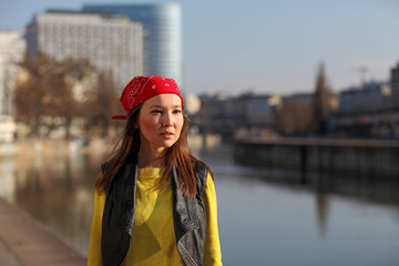 Beautiful Asian woman in yellow sweater and with red headscarf standing at a river in a European city