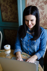 Fototapeta na wymiar Beautiful happy young woman dressed in denim shirt sitting in the cafe working at a laptop drinking coffee