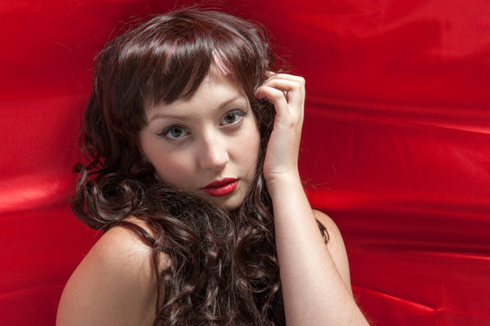 young lady with long brunette curled hair laying on red satin cloth