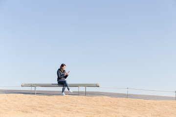 Woman using cellphone and sitting at bench with blue sky