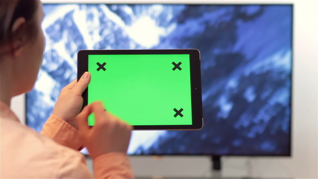 Woman watches television while holding and tapping on a tablet computer device with green screen.