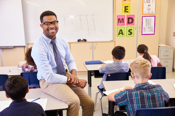 Smiling elementary school teacher sitting on a pupil’s desk