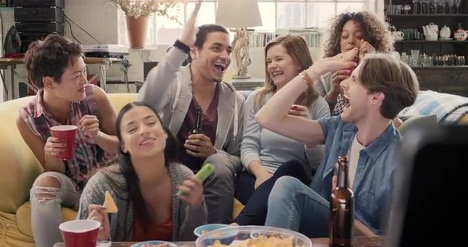 Diverse group of student friends arms raised celebrating goal watching sports event on TV together eating snacks  drinking beer 