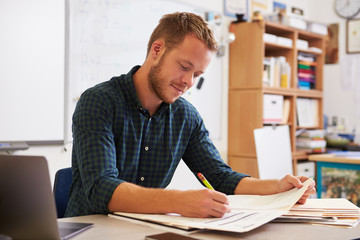 Young bearded male teacher at desk marking students’ work