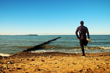Tall man in black suit exercising on stony beach at breakwater.