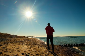 Runner  in red sportswer running and exercising on stony beach at breakwater. - obrazy, fototapety, plakaty