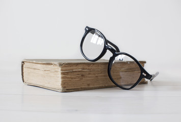 Vintage book on white wooden background with black reading glasses on top