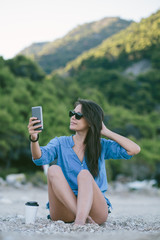 Portrait of young woman sitting at the beach and making selfie