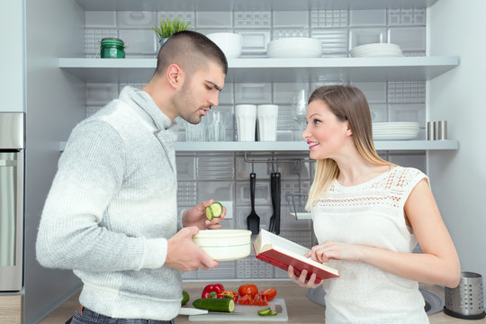 Happy young couple is preparing healthy meal together in the kitchen. Young woman is reading recipe from the cookbook, and young man is trying to follow her instructions.