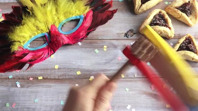Hand play with Traditional wooden Purim gragger over Purim Jewish holiday food and objects: Hamantasche and carnival mask.