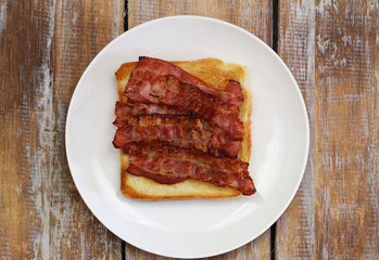 Crispy bacon on toast on white plate on rustic wooden surface
