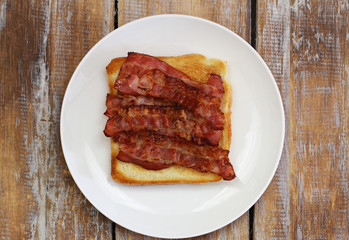 Crispy bacon on toast on white plate on rustic wooden surface
