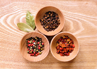 Three wooden bowls with pepper, cloves and slices of dried peppers on a light wooden table. Next to the bowl are two of dried bay leaves.