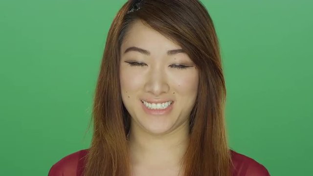 Young Asian woman awkwardly smiling, on a green screen studio background 