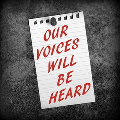 The message Our Voices Will Be Heard in red text on a scrap of lined paper pinned to a grunge background in black and white for effect