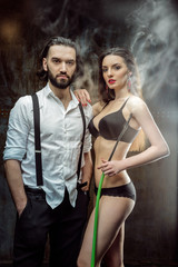 Handsome bearded man in white shirt with sexy young brunette woman in black underwear smoking hookah with weathered metal wall on background