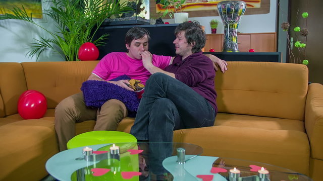 Gay couple seating on a sofa feeding etch other with cookies in a living room, footage taken in slow motion.
