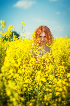 Smiling red-haired pretty girl looks out of the yellow flowers on the field
