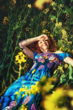 Happy red-haired young girl in a blue dress, lying in a green field with yellow flowers
