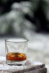 Whiskey sitting on a wood table with ice snow pellets. In background snow covered trees are in soft focus. Lots of copy space.