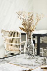 Dried flower in transparent vase on marble table with blurry background of table and chair