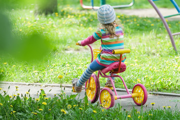 Back view full length portrait of preschooler girl riding kids pink and yellow tricycle on playground track