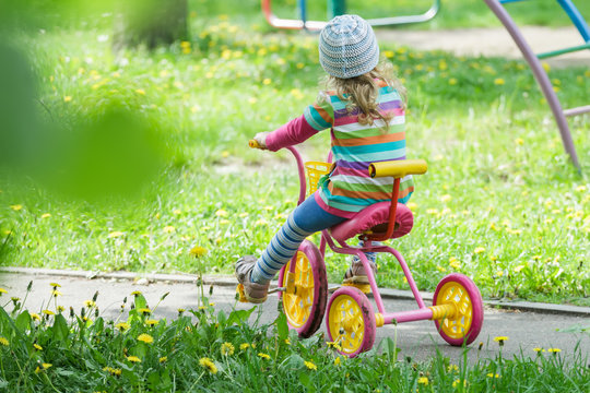 Back View Full Length Portrait Of Preschooler Girl Riding Kids Pink And Yellow Tricycle On Playground Track