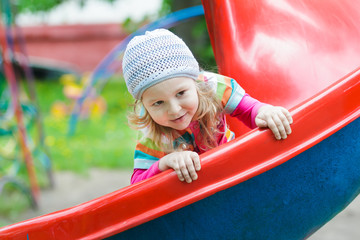 Fototapeta na wymiar Smiling little blonde girl sliding down red plastic playground slide outdoors on spring