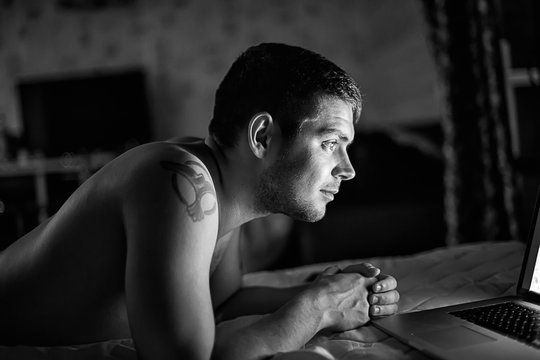 young man with naked torso on the bed with laptop/ black and white photo