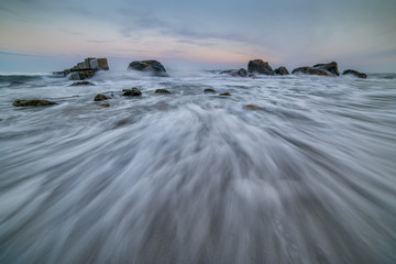 Gentle calm seascape. In the background light teploho sun at dawn, dusk. Long white stripes on the sand by the waves downwind. Rocks, waves crashing on the rocks, mist from the spray, the nebula.
