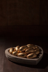 Close-up of Easter eggs in gold foil in a heart shaped wooden bowl against a dark background. 