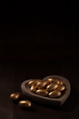 Close-up of Easter eggs in gold foil in a heart shaped wooden bowl against a dark background. Focus is on the eggs in the foreground.