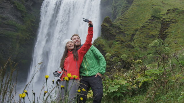 Selfie couple taking smart phone picture of waterfall outdoors in front of Skogafoss on Iceland. Couple visiting famous tourist attractions and landmarks in Icelandic nature landscape on the ring road