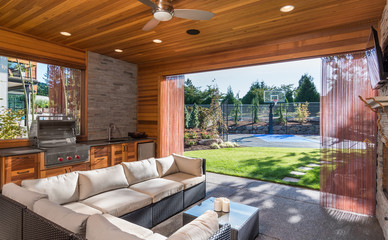 Beautiful covered patio outside new luxury home with television, fireplace, and lush green yard leading to basketball court