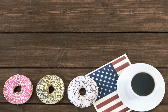 Four American donuts are lying in a row on a wooden table. Cup of coffee is lying on the napkin in american flag design.