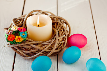 Easter background with candle in a nest of twigs and colored eggs on white wooden background