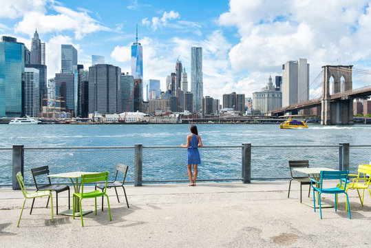 New York City Skyline Waterfront Lifestyle - American People Walking Enjoying View Of Manhattan Over The Hudson River From The Brooklyn Side. NYC Cityscape With A Public Boardwalk With Tables.