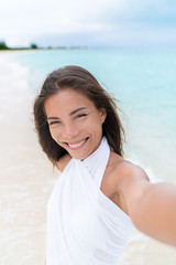 Fototapeta na wymiar Selfie of beautiful Asian chinese caucasian mixed race woman on beach wearing white cover-up dress. Pretty young adult holding camera phone smiling during summer vacation caribbean travel.