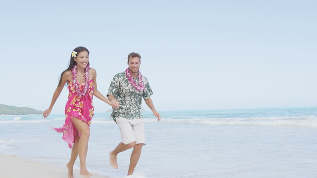 Cheerful couple in Aloha clothing running on Hawaiian beach. Man and woman are holding hands while spending leisure time on sea shore. Visitors are enjoying against sky on sunny day.