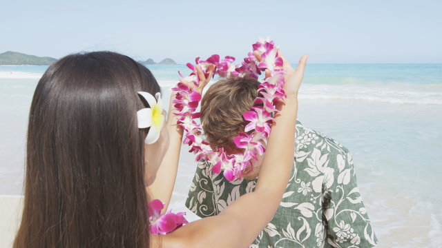 Young woman putting lei garland of pink orchids around man's neck at Hawaiian beach. Handsome male is in Aloha clothing. Happy couple is enjoying their summer vacation.