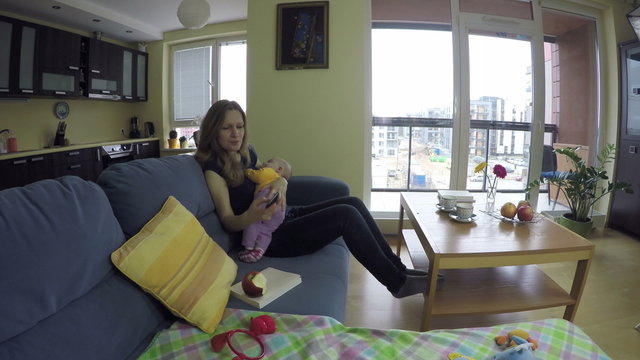 Mother eat apple fruit with cute sleeping baby girl on her hands. Woman answer mobile phone and talk quietly with smile. Wide angle shot.

