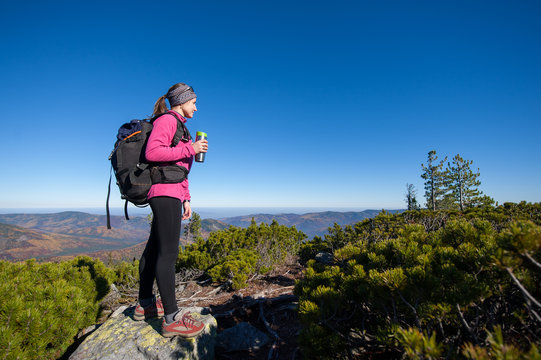 Young woman backpacker reaching her goal standing on peak of the mountain, holding cup of coffee and enjoying the beautiful landscape. Sunny autumn time.