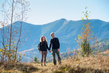 Young happy couple tourists holding hands smiling and looking into distance on a warm sunny autumn day in the mountains. Mountain on the background.