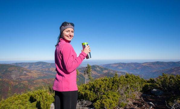 Young lady hiker smiling holding the thermos and looking at the camera. Happy woman enjoying beautiful day up on the mountain. Colorful fall time.