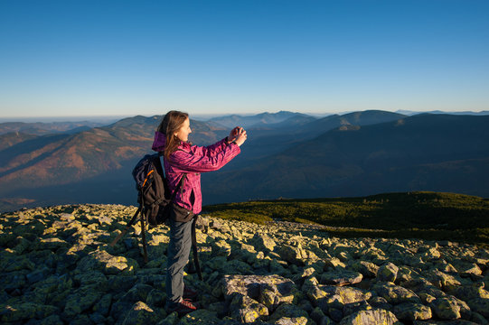 Young beautiful woman standing on rocky mountain top and taking picture with her smartphone. Beautiful mountains on the background. Healthy lifestyle concept. Sunny fall evening.