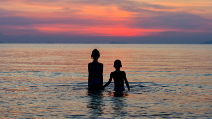 Silhouettes of girl and boy standing in the sea holding hands and watching tropical sunset
