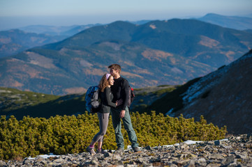 Naklejka premium Szczęśliwa młoda para stoi cieszyć się przerwą podczas wędrówki na wakacjach, uśmiechając się i przytulając. Wielkie piękne góry w tle. Ciepły i słoneczny jesienny dzień.